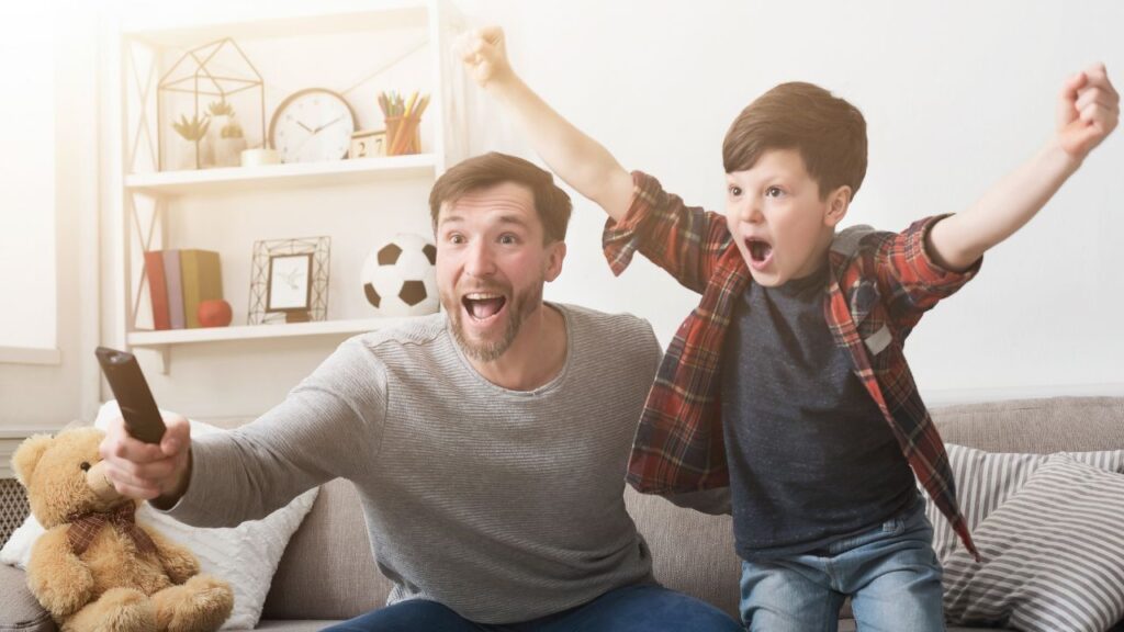 pai e filho comemorando enquanto assistem partida de futebol